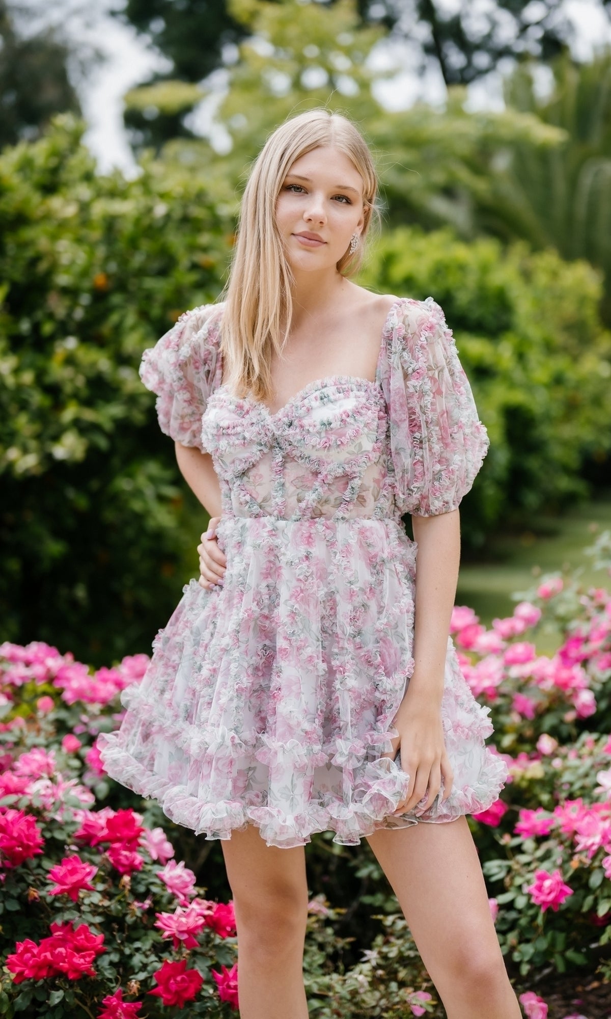 A young woman wearing the Aleta 1304 Short Sleeve Short Print Prom Dress stands in a garden surrounded by pink flowers and lush greenery.
